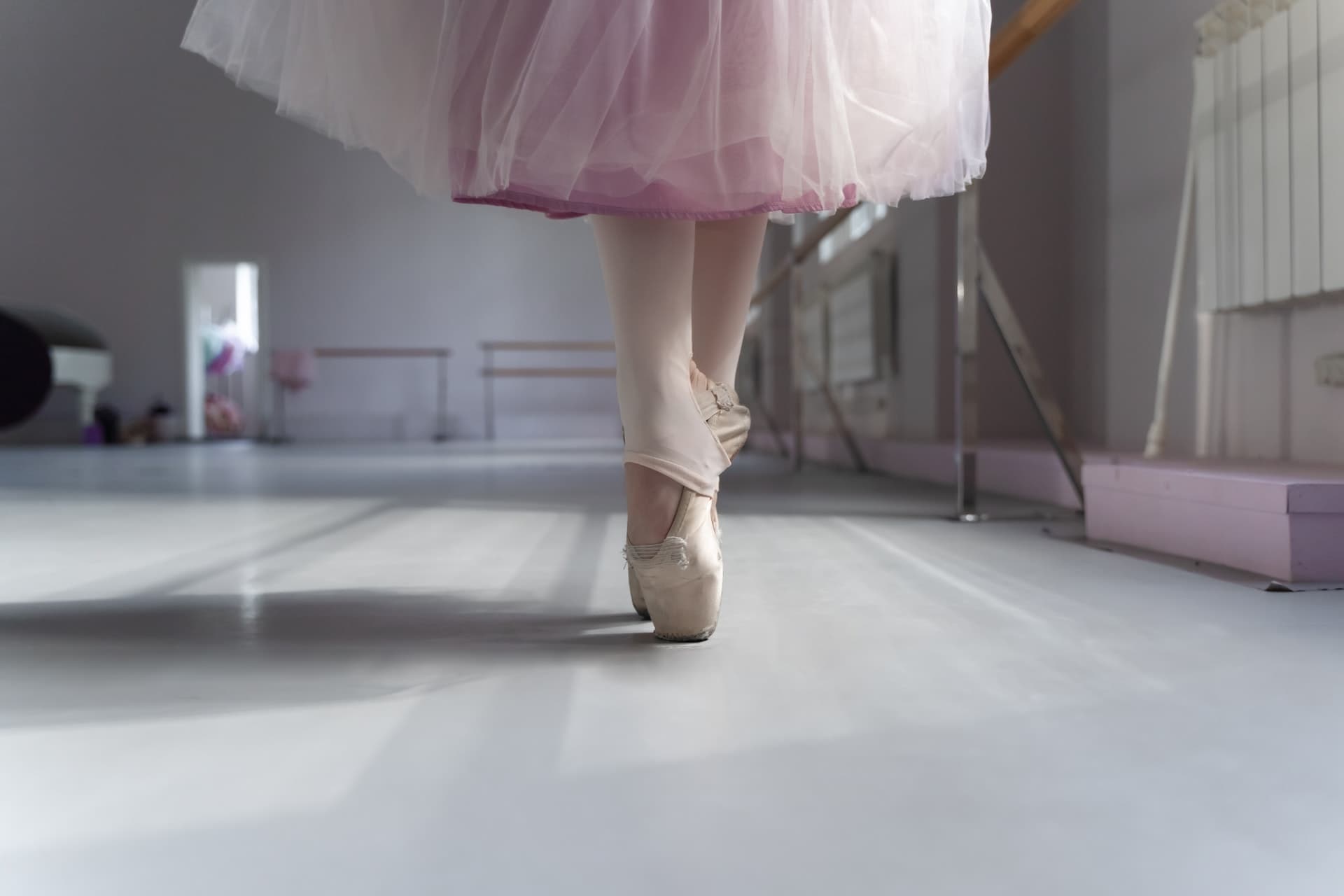 the feet and lower legs of a female ballet dancer on her toes in very light pink ballet shoes at a dance studio, she has a pink skirt and the ballet dance studio is empty except for her