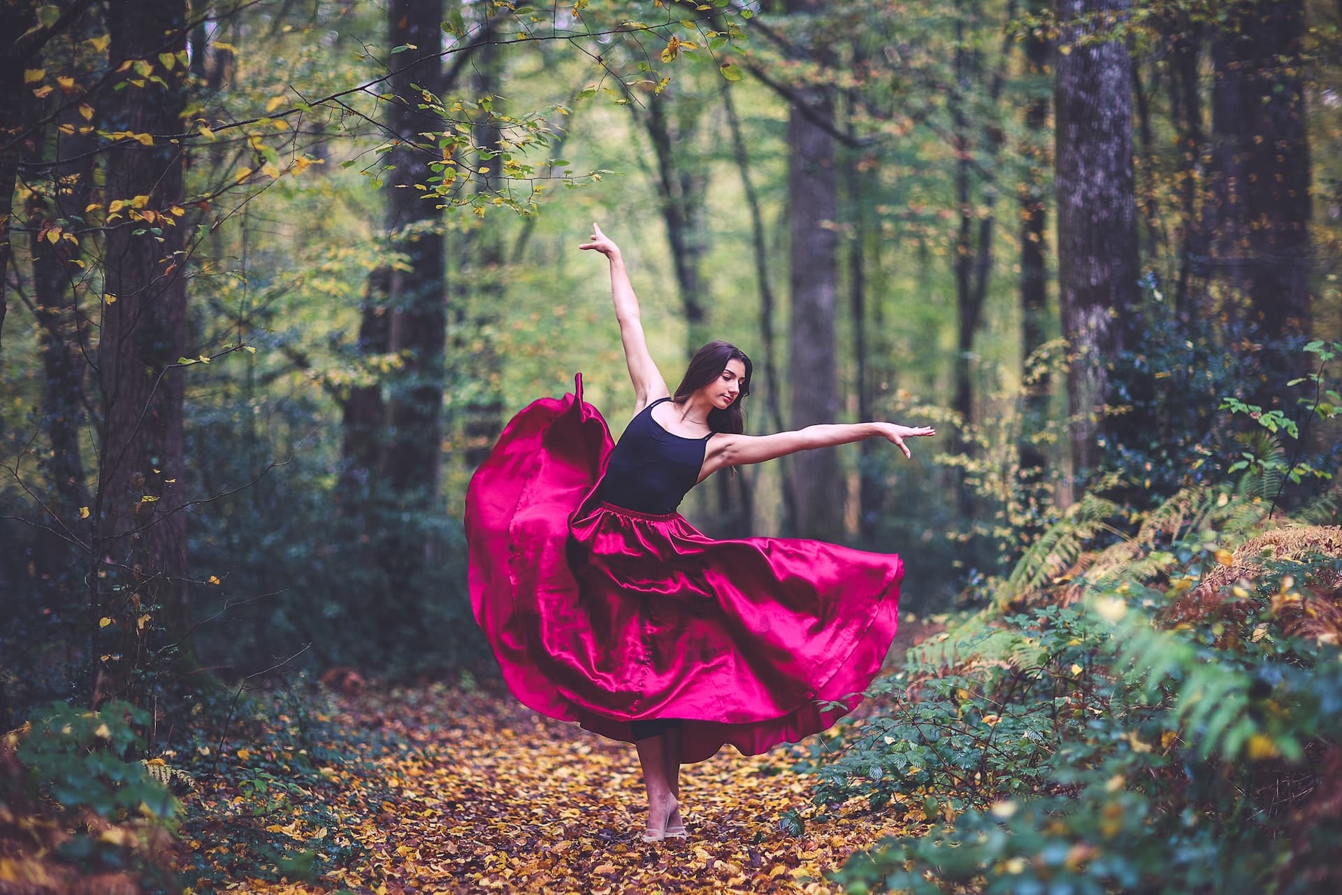 A young modern ballet dancer in a striking fuscia skirt and black top is posing dramatically in a classis ballet pose, she is outdoors in nature surrounded by a forest