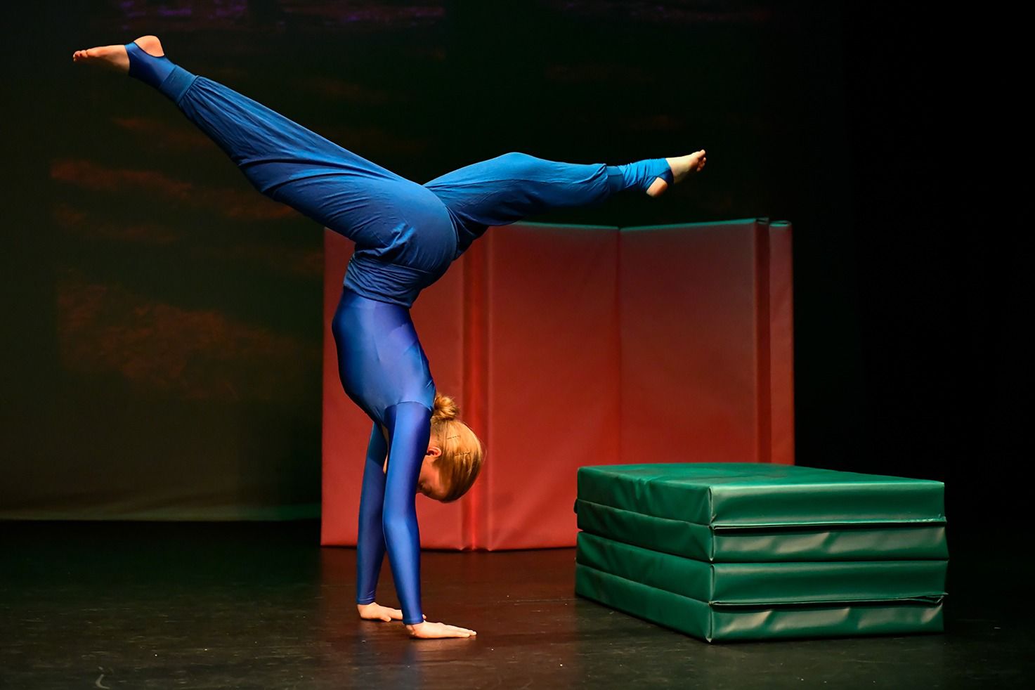 A teenage female student Acro ballet dance at Ruth Shine School of Dance performs dramatically on stage, she is in a hand-stand position with her legs split front to back and she is wearing a blue outfit with long sleeve