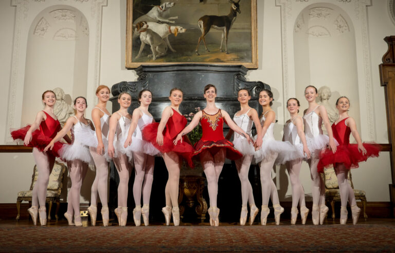 Ballet dance teacher Ruth Shine in a red ballet dress poses with eleven of her young female ballet students in a beautiful and elegant Georgian room