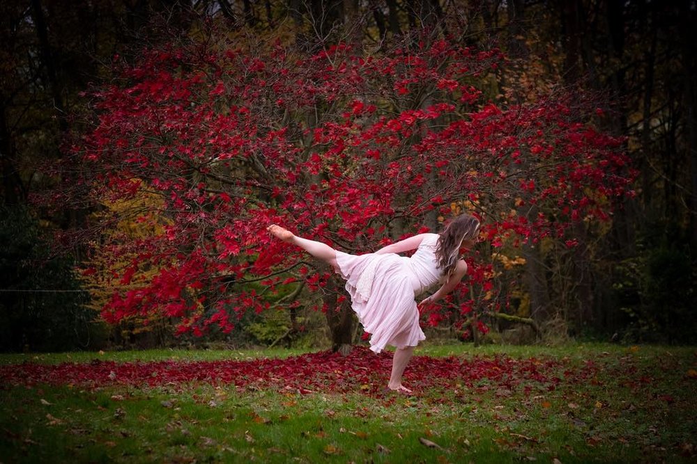 an adult female contemporary dance student poses in a dramatic stance while barefoot. It is autumn and in the background a red maple tree is dropping leaves on the grass