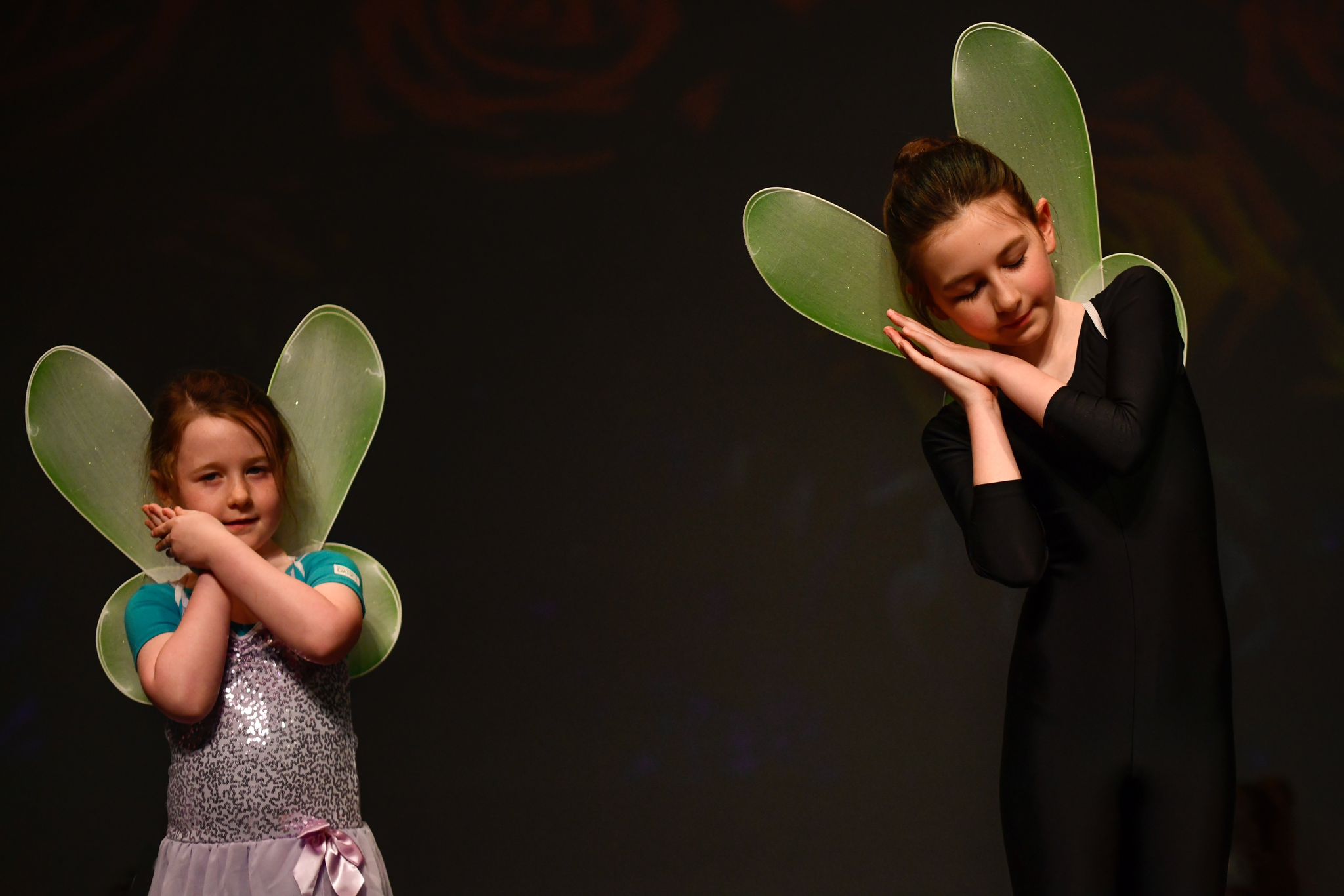 two young Irish girls wearing fairy wings on their leotards make a gesture of sleeping with their hands, they are on stage performing their ballet dance routine at a performance night for parents in Dublin, Ireland
