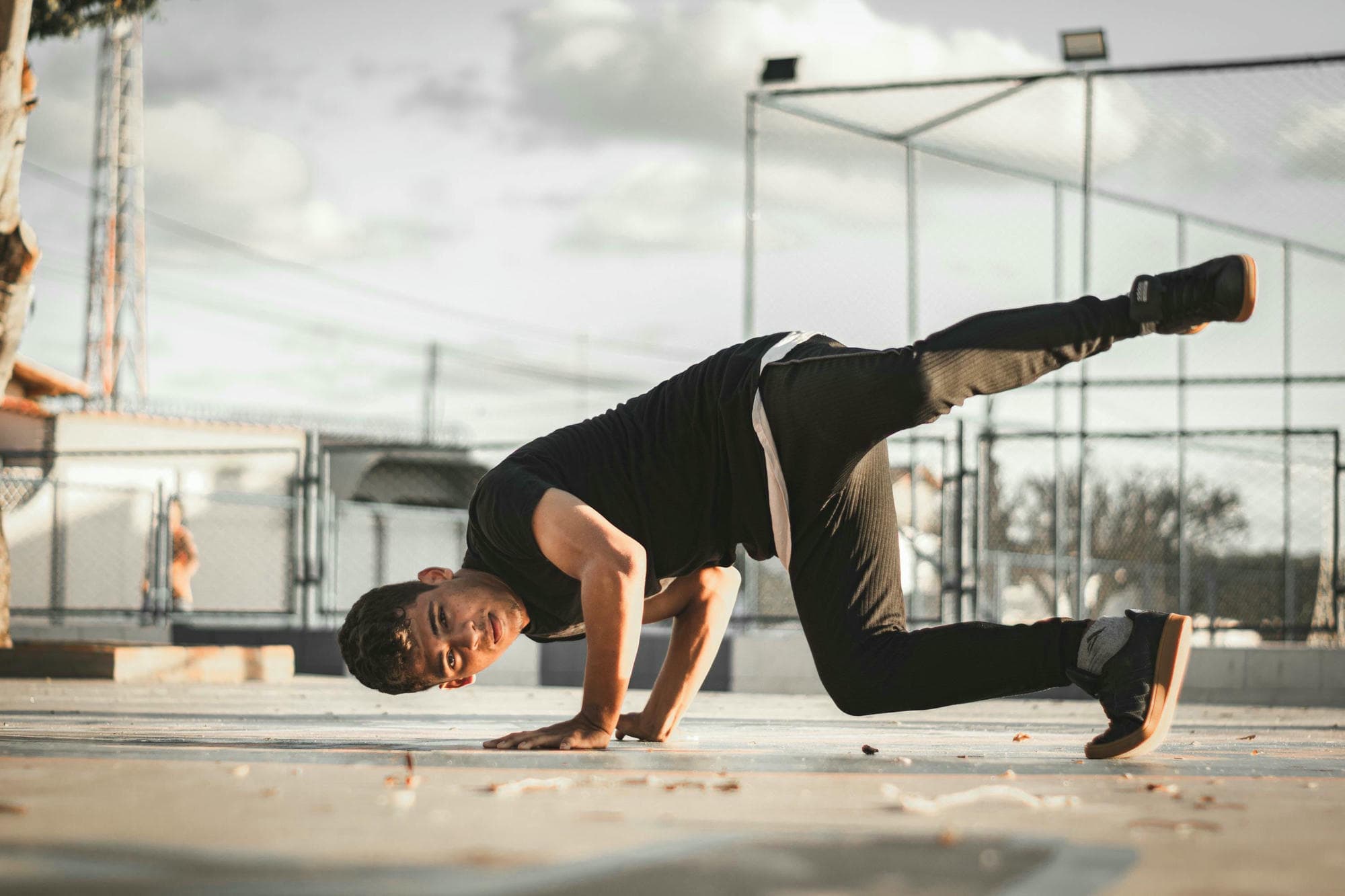 A male breakdancer in black , performing an acrobatic pose on the ground at a sunny outdoor gym with a metal fence and roof in the background, in the style of Unsplash photography with natural light, pastel colors, and a minimalistic aesthetic