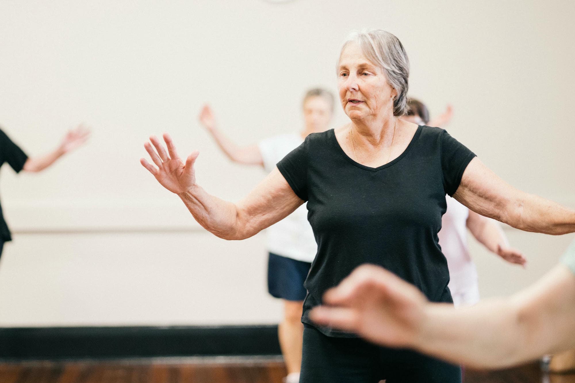 A photograph of an elderly woman leading her group in dance, standing with open arms and a focused expression against the backdrop of another person dancing. The studio is a large, white room with soft lighting highlighting their movements. In the front view, the focus is on the older women engaging in various dance poses, embodying energy and movement