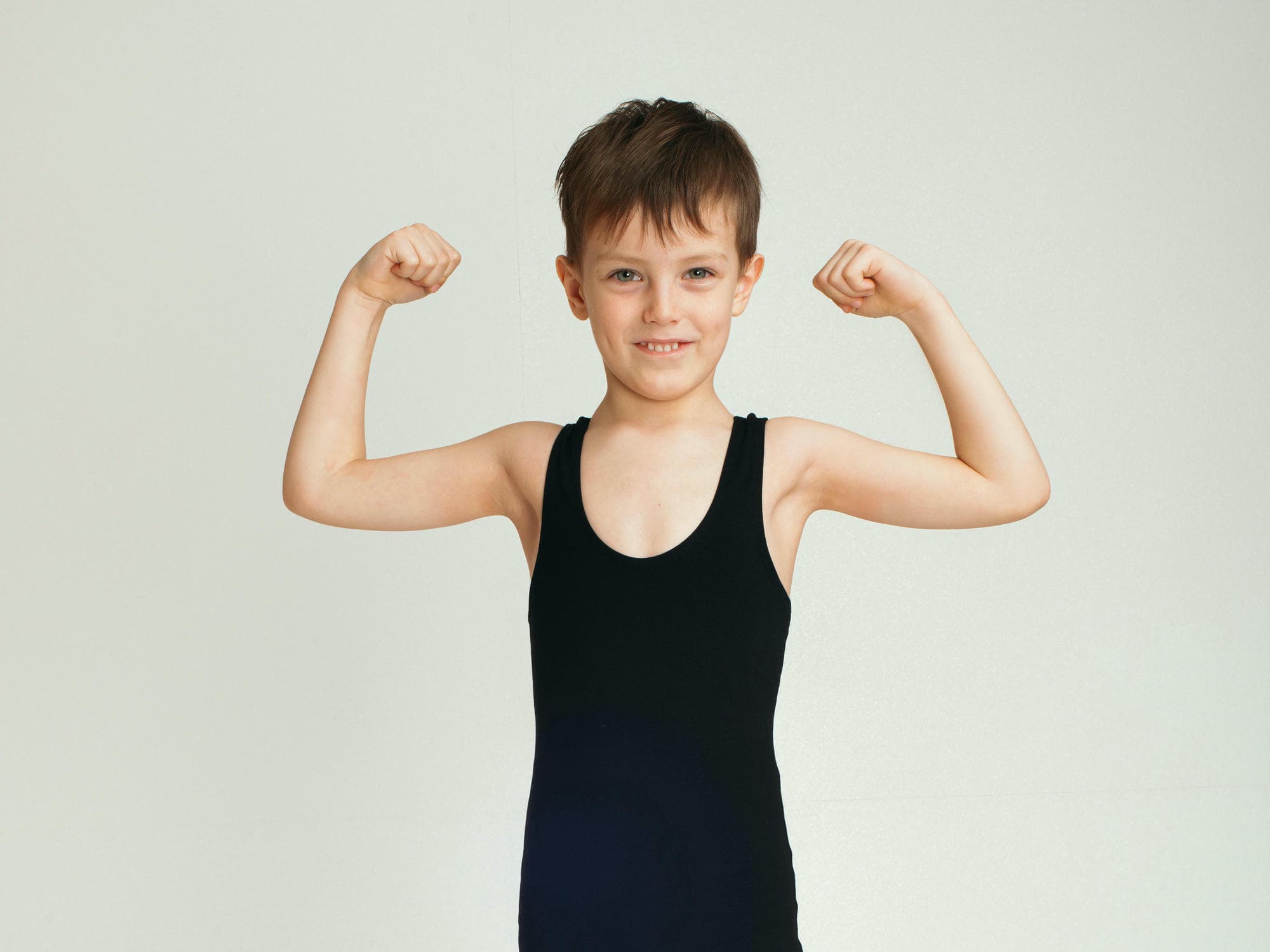 Photo of a tall, skinny young boy, a ballet dancer, in black leotards, smiling and flexing his biceps to show off his muscles against a white background